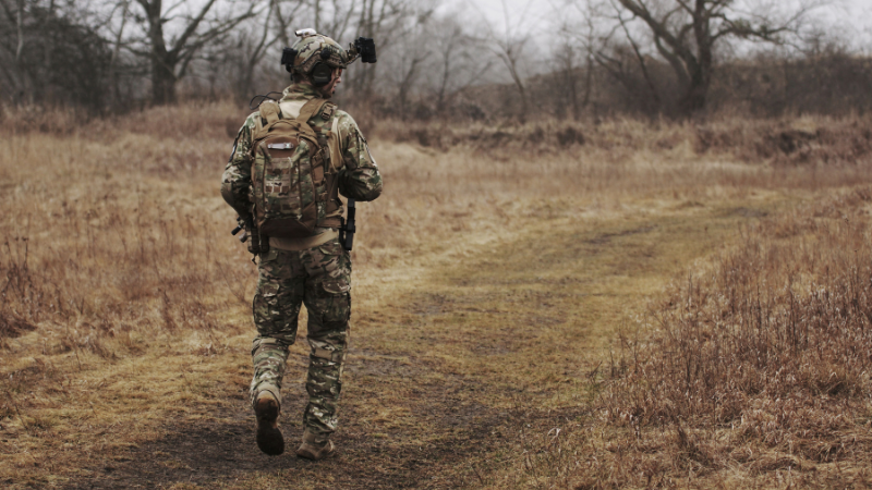 A soldier in camouflage walking along a grassy path through a forested area.
